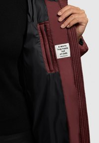 Innenseite einer kastanienbraunen und schwarzen Jacke mit gestepptem Muster, die ein Stoffetikett trägt, auf dem steht: „Eine warme Umarmung aus Stoff.“