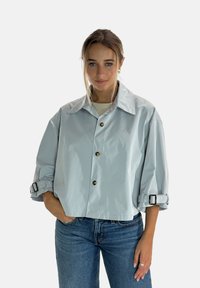 Lyseblå kort jakke lavet af glat stof, med en bred krave, store knapper og opkørte ærmer med spænde-detaljer.