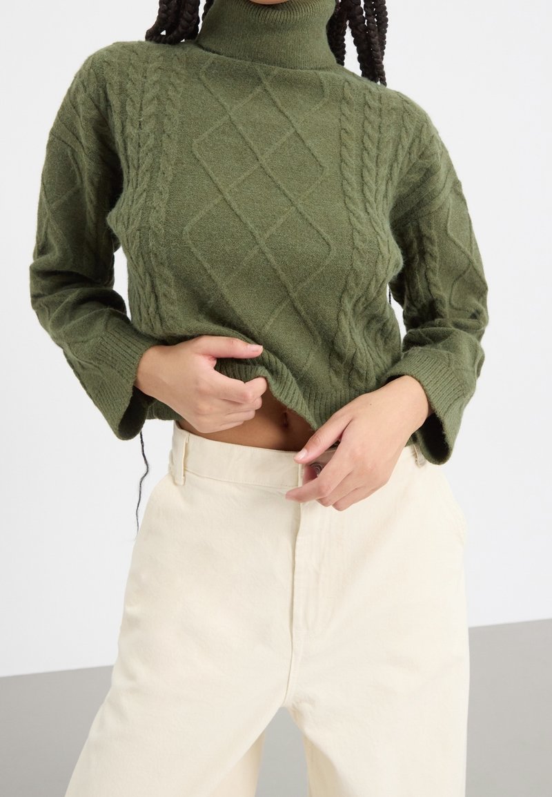 Personne portant un pull à col roulé en maille torsadée vert olive et un pantalon crème taille haute, tenant l'ourlet du pull pour dévoiler le ventre.