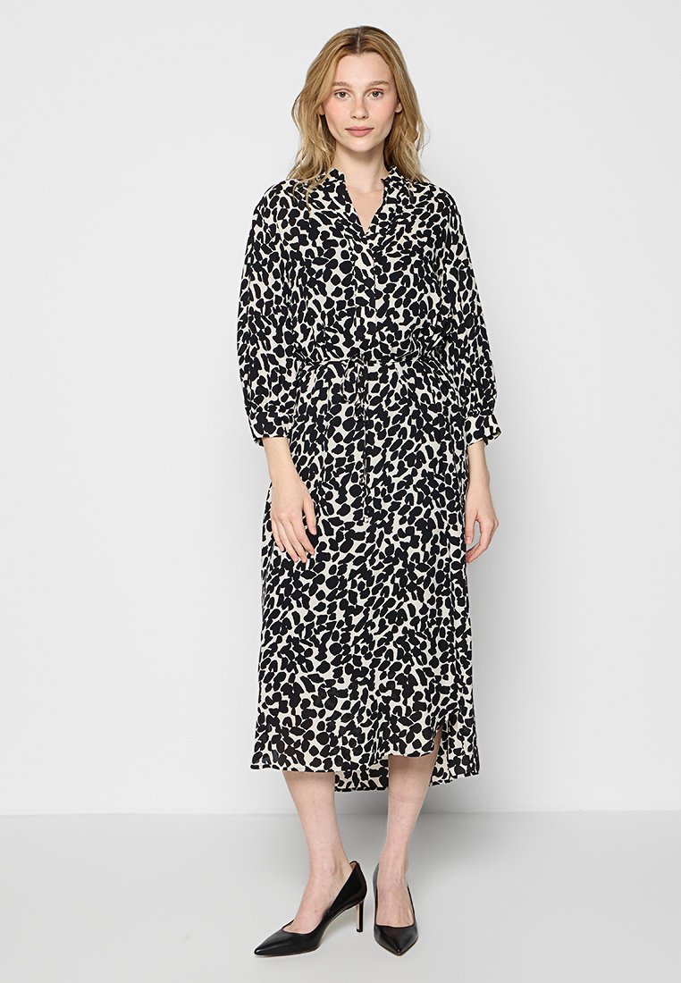 Langärmliges, knielanges Kleid mit einem schwarz-cremefarbenen abstrakten Muster, Taillenband und leicht ausgestellt am Saum, kombiniert mit schwarzen High Heels.