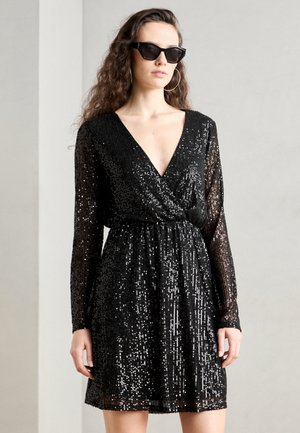 Robe noire en sequins avec un décolleté en V profond et des manches longues, cintrée à la taille. Le tissu présente une finition chatoyante et texturée.