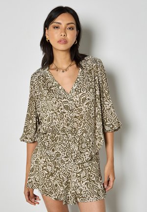 Kobieta ubrana w oliwkowo-beżową bluzkę z dekoltem w serek z wzorem oraz dopasowane szorty, do tego złote kolczyki koła i delikatny naszyjnik.