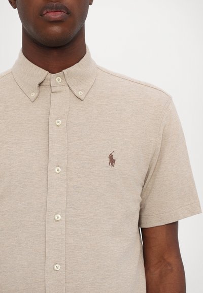 Camisa polo de manga corta beige, fabricada en tejido texturizado, con cuello abotonado y un pequeño logo bordado marrón en el lado izquierdo del pecho.