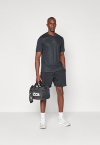 T-shirt de sport noir avec un motif texturé, associé à un short noir. Porte un sac de sport noir avec une marque blanche. Baskets et chaussettes blanches.