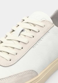 White leather sneaker with light beige suede accents, textured surface, flat cream laces, and a subtle logo embossed on the side.