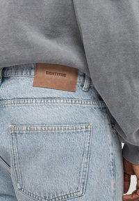 Ljusblåjeans med en läderlapp märkt "EIGHTYFIVE" på baksidan, ihopparat med en grå sweatshirt som delvis täcker midjebandet.