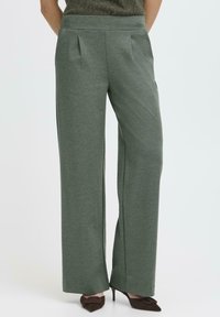 Pantaloni evazați dintr-un material verde închis, cu pliuri frontale, textură netedă și buzunare laterale, asortați cu tocuri ascuțite maro.