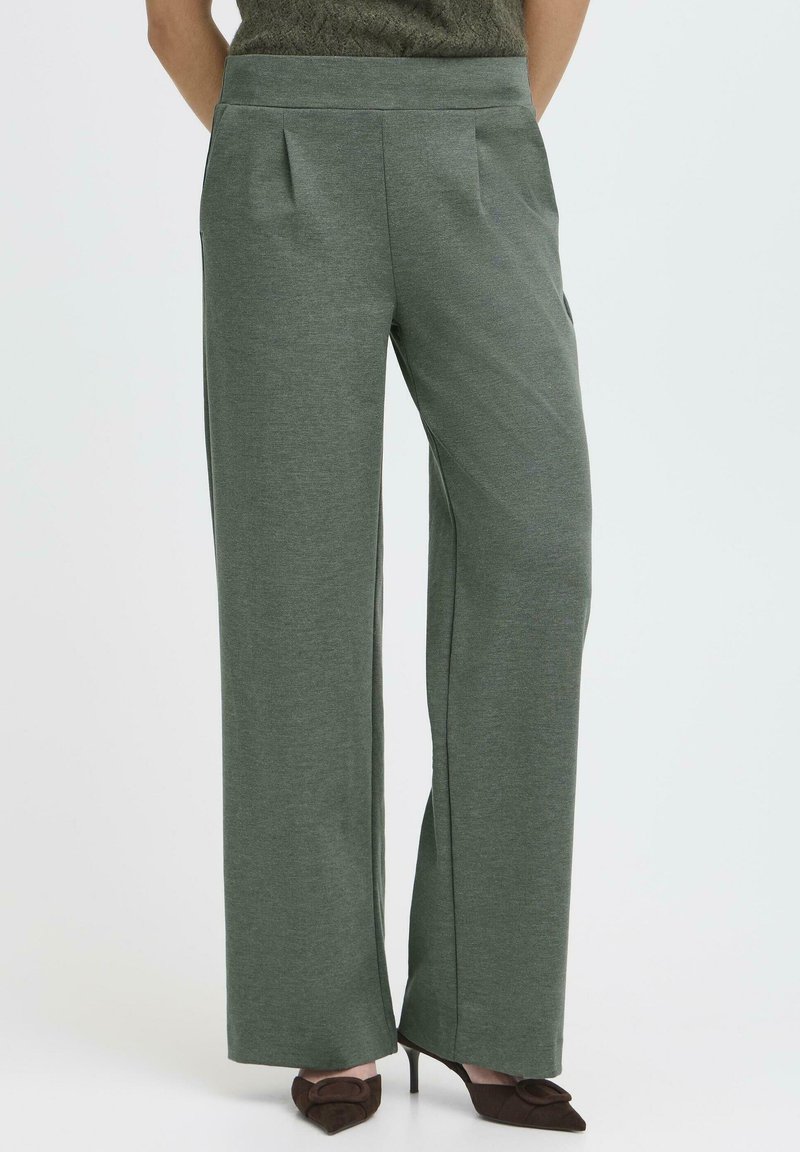 Pantaloni evazați dintr-un material verde închis, cu pliuri frontale, textură netedă și buzunare laterale, asortați cu tocuri ascuțite maro.