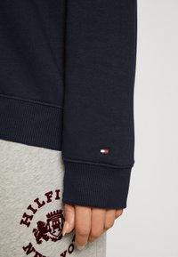 Felpa blu navy con polsini a coste, dotata di un piccolo logo Tommy Hilfiger. Pantaloni della tuta grigi con un dettagli grafico ricamato di color rosso scuro.