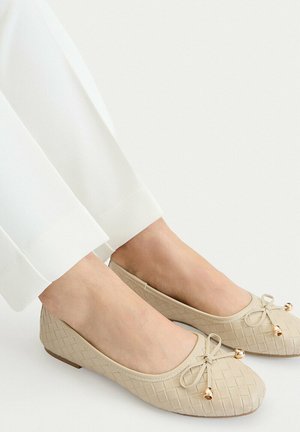 Bailarinas beige de tejido con pequeños lazos decorativos y campanas doradas, usadas con pantalones blancos y pies descalzos.