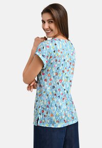 Helles, kurzärmliges Oberteil in Blau mit einem mehrfarbigen, abstrakten Muster. Mit Rundhalsausschnitt und Seitenschlitzen. Weicher, strukturierter Stoff.