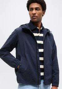 Ung mand iført en marineblå hættejakke over en marineblå og hvidstribet poloshirt med lyseblå jeans, der kigger til siden.