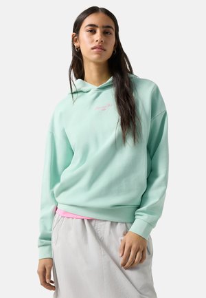 Jonge vrouw met lang donker haar die een mintgroene hoodie en een lichtgrijze rok draagt, staand tegen een effen achtergrond.