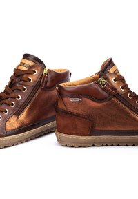 Bottines en cuir marron avec une finition lisse, dotées d'une fermeture éclair sur le côté, de lacets ronds et d'une semelle en caoutchouc texturé. Logo de la marque visible sur le côté.