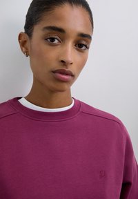 Mujer con el cabello liso recogido hacia atrás, llevando aretes pequeños de aro y una camisa blanca con capas debajo de un suéter de cuello redondo morado.