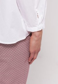 Bras droit portant une chemise blanche à manches longues avec un poignet boutonné reposant à côté d'un pantalon rouge et blanc à motifs.