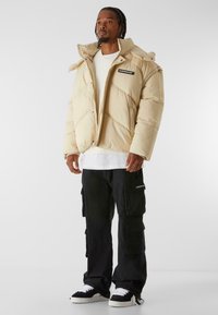 Beige puffer jacket with a high collar, zip closure, and diagonal stitching detail. Paired with black cargo pants and sneakers.