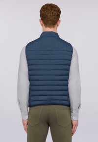 Námořnická modrá prošívaná vesta s vertikálním prošíváním a stojáčkovým límcem. Nositá přes světle šedou košili a dobře padnoucí olivové kalhoty.