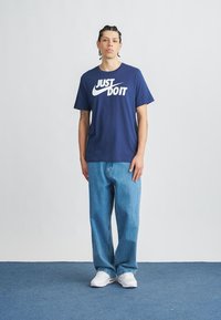 Μπλε βαμβακερό μπλουζάκι με λευκό λογότυπο "Just Do It", συνδυασμένο με ανοιχτόχρωμα τζιν και λευκά αθλητικά παπούτσια. Το μοντέλο στέκεται μπροστά σε ανοιχτό φόντο.