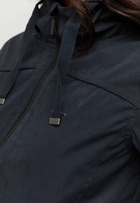Ciemnoniebieska kurtka z zamkiem błyskawicznym z przodu, regulowanym kapturem ze sznureczkiem i subtelną fakturą. Posiada metalowe elementy i szczegóły szwów.