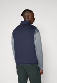 Gilet in pile blu navy con maniche lunghe grigie, colletto alto e texture liscia, indossato sopra pantaloni cargo verdi, visto da dietro.