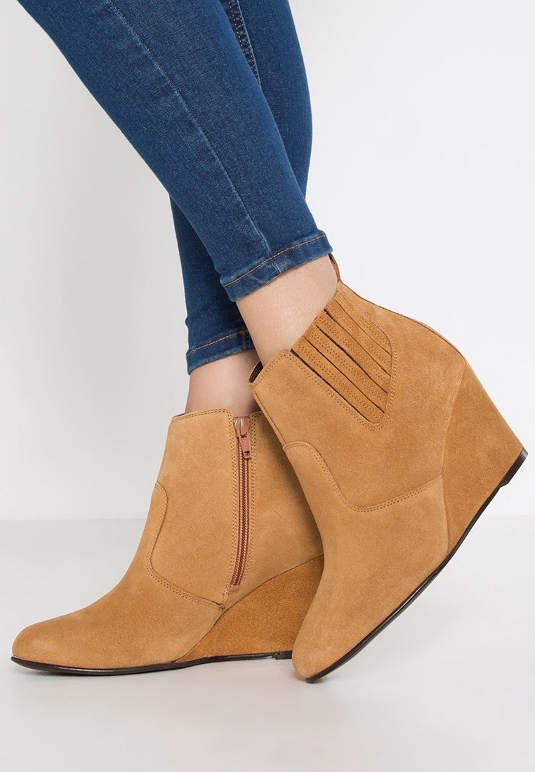 Bottes d cheville en daim beige avec un talon compens, dotes d une fermeture glissire latrale et d accents c ts nervurs verticalement sur la tige. Texture lisse et bout pointu.