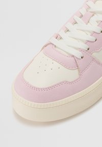 White and pink sneakers with a suede upper, textured rubber sole, perforated toe, and flat white laces. Rounded shape with contrasting accents.