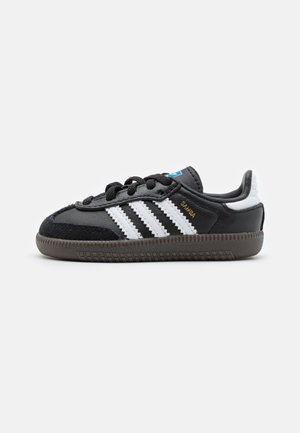 Baskets Adidas Samba noires avec des bandes blanches, des lacets noirs, une semelle en caoutchouc, et un détail blanc au talon sur fond blanc uni.