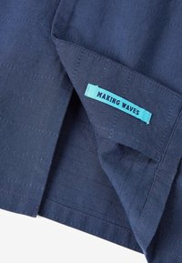 Tissu bleu marine avec des rayures texturées subtiles. Comprend une étiquette bleu clair portant la mention "MAKING WAVES" cousue sur l'ourlet. Bords propres et soignés.