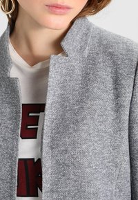 Grijze textuurjack met een hoge kraag, gemaakt van een zachte stof en met strakke lijnen, gelaagd over een wit shirt met rode letters.