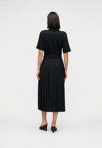 Μαύρο πλισέ μακρύ φόρεμα με κοντά μανίκια και ζώνη. Λείο ύφασμα με δομημένη σιλουέτα, συνδυασμένο με μαύρα mule πέδιλα με τακούνι.