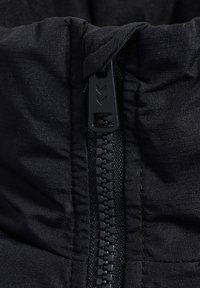 Sort jakke tæt på med en tekstureret stof, en central lynlås og trækflig, der viser et mærkelogo på lynlåstrækket.