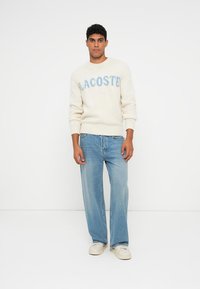 Pulover tricotat crem cu text albastru "LACOSTE", asortat cu blugi drepti de culoare bleu deschis și adidași bej. Design simplu cu manșete ribotate.