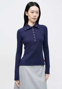 Tricou polo cu mâneci lungi, de culoare bleumarin, cu guler structurat, fante decorative cu nasturi și textură subtilă nervurată. Etichetă roșie la tiv.