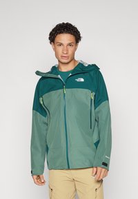 Veste imperméable verte et teal avec fermeture éclair à l'avant et capuche ajustable. Dotée de deux poches zippées et d'un tissu texturé. Design léger.