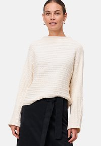 Femme portant un pull écru en maille torsadée à manches longues et une jupe noire avec une ceinture nouée, se tenant devant un fond uni.