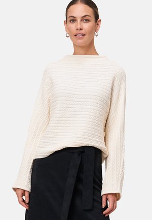 Femme portant un pull écru en maille torsadée à manches longues et une jupe noire avec une ceinture nouée, se tenant devant un fond uni.
