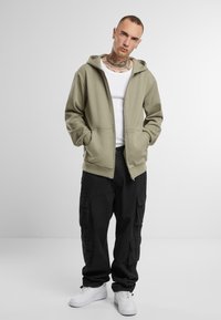 Groene zip-up hoodie met zakken, gedragen over een witte tanktop, gecombineerd met zwarte cargobroek en witte sneakers. Glad textuur van de stof.