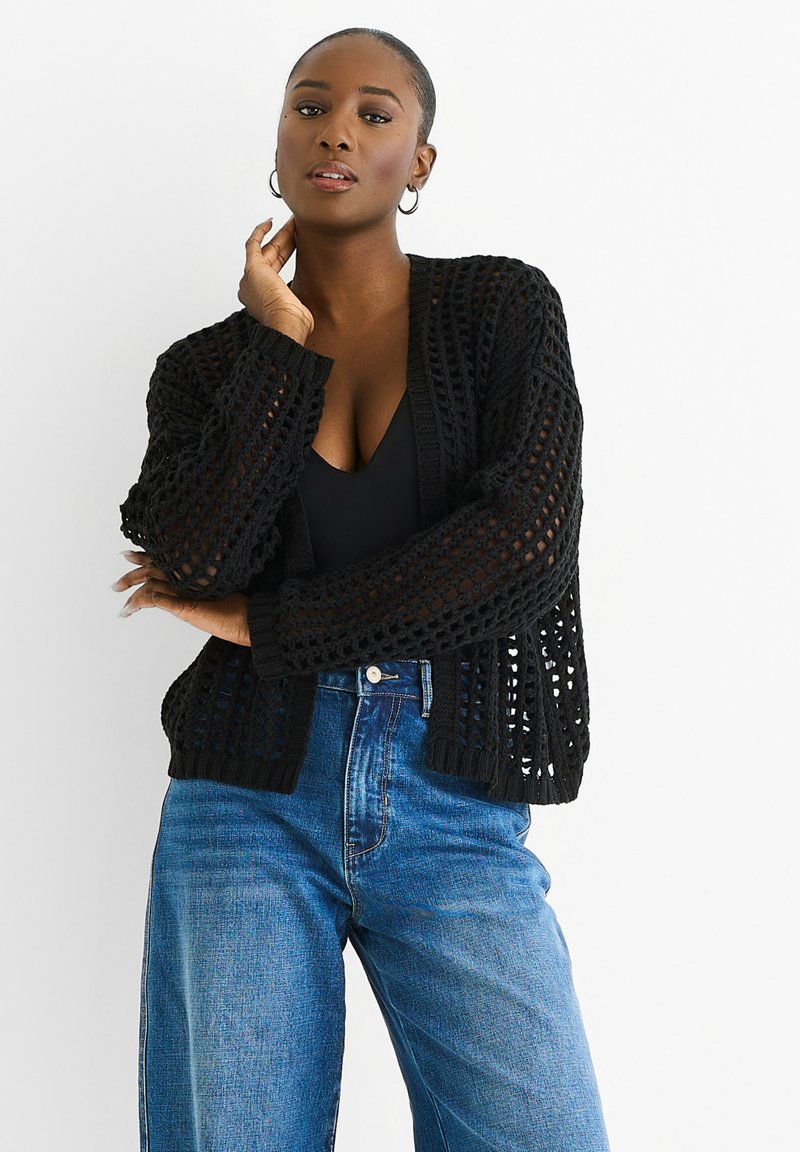 Black, open-knit cardigan with long sleeves, loose fit, and ribbed cuffs. Paired with high-waisted light blue denim jeans.