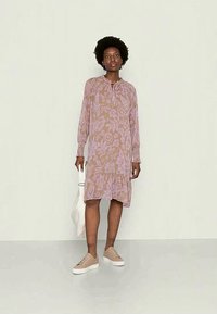 Vestido de manga longa com estampa floral em tons de rosa claro e bege, apresentando um decote redondo com detalhe de laço e uma bainha larga. Usado com ténis.