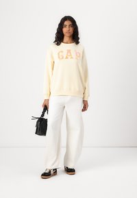 Krem sweatshirt med "GAP"-logo i pastellfarger, kombinert med hvite vide bukser og svarte joggesko, holder en liten svart håndveske.