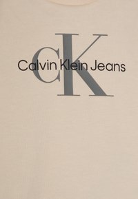 Beige puuvillainen t-paita, jossa suuri musta "Calvin Klein" -teksti ja harmaa "CK"-logo. Pyöreä pääntie resorireunuksella, sileä pinta.