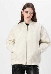 Vaaleanbeige bomber-takki, vetoketjukiinnitys, ribbaatut hihansuut ja helma, minimibrändäys. Yhdistetty kiiltäviin mustiin housuihin. Suora kangaslaatu.