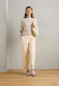 Cream ribbed sweater, light blue turtleneck, loose cream pants, gray sandals. Model holding a small beige bag, standing on wooden floor.