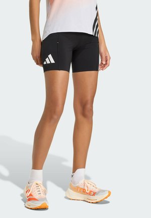 Osoba nosící černé cyklistické šortky Adidas, bílé kotníkové ponožky a bílo-oranžové běžecké boty Adidas Terrex, stojící na bílém pozadí.