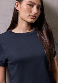 Giovane donna con lunghi capelli lisci, che indossa orecchini a cerchio dorati e una maglietta blu scuro, in piedi contro un muro di texture chiara.