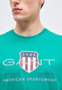 Νεαρός άνδρας που φοράει πράσινο μπλουζάκι GANT με λογότυπο ασπίδας σε κόκκινο, λευκό και μπλε χρώμα και το κείμενο «1949 American Sportswear» μπροστά.