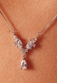 Collar en tono plateado que presenta cristales claros en forma de pera y redondos dispuestos en forma de V, con una piedra colgante más grande en el centro.