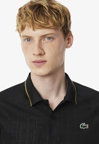 Svart polo skjorta med geometriskt mönster, med en grön och vit logotyp, liten krage, och gul piping längs nacklinjen.