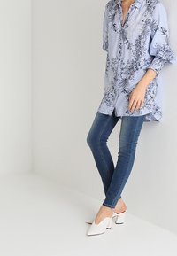 Vaaleansininen nappikauluspaita, jossa on kukkakuosi, yhdistetty tummansiniset skinny-farkut ja valkoiset avonaiset mules-kengät. Rentoa asua kerroksilla.